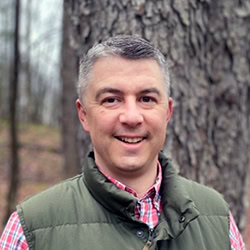 Thad Taylor smiles outdoors in front of tree wearing red plaid shirt and green vest