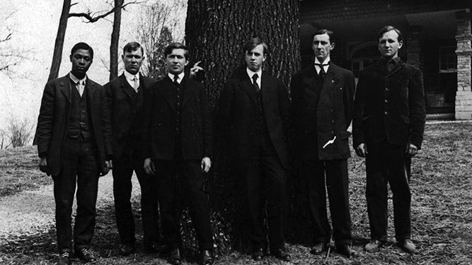 Old black and white photo featuring six young men in dark suits and ties standing in front of a large tree