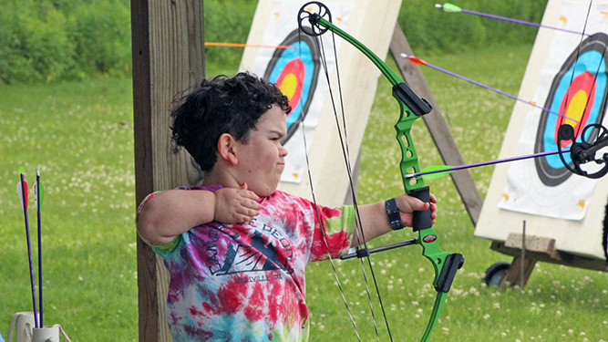 An indivdual uses a bow to shoot towards archery targets. Arrows and targets appear in the background.