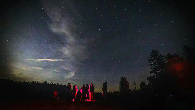 Group of people in the distance illuminated by red lighting underneath dark sky and stars at Cherry Springs State Park.