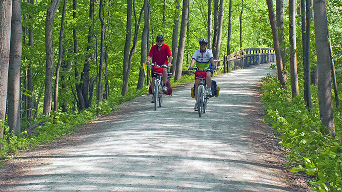 Two people ride bikes on flat gravel trail through dense trees and green foliage in Ohiopyle State Park.
