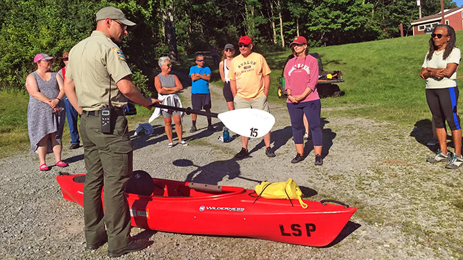 A park ranger demonstrating kayak paddling to a group of 8 individuals. Trees and buildings appear in the background.