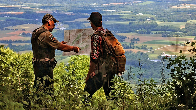 Forest Ranger Steve Shaffer and Intern Connor Duncan reading a map at while overlooking various trees, fields, and foliage.
