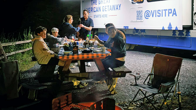 Governor Josh Shapiro and family at a picnic table at night next to Recreational Vehicle at a campsite at Lyman Run State Park.