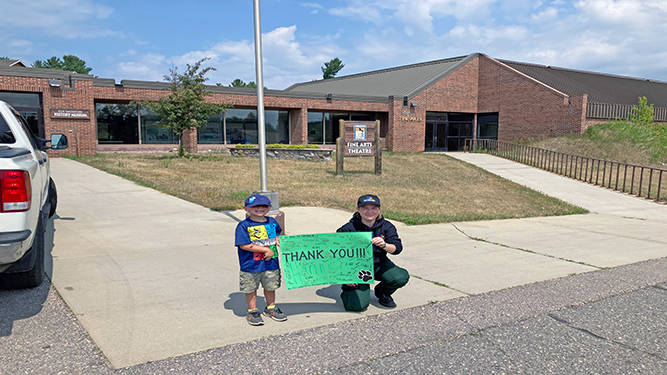 A person and a child hold a Thank You sign in front of a Fine Arts Theatre.