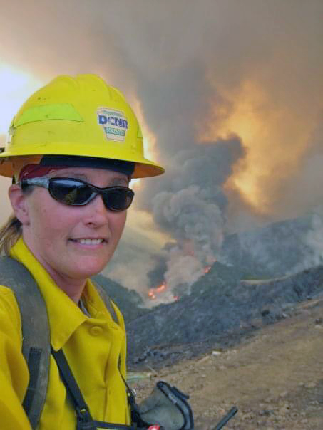 A firefighter stands in front of a distant wildfire.