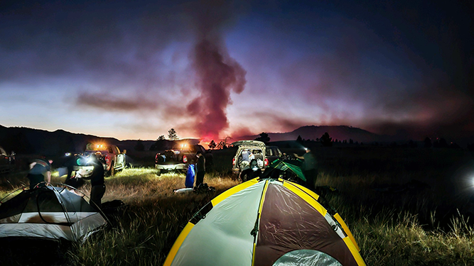 A firefighting crew stays at a campsite with a wildfire in the distance.