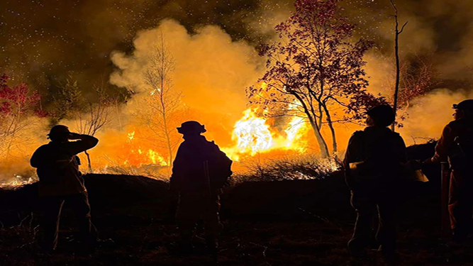 Four firefighters gather with a wildfire in the distance.