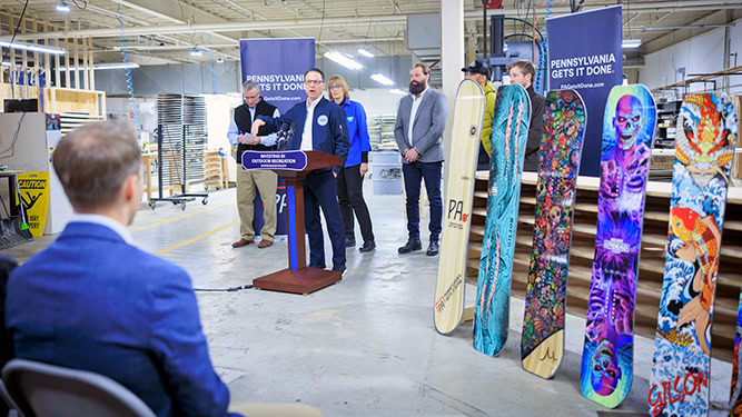 Governor Josh Shapiro speaks at a podium at a outdoor snow company warehouse. Colorful snowboards appear to his side.